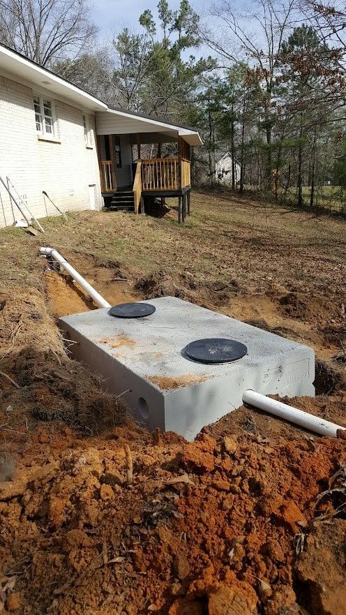 A rectangular concrete septic tank in an excavated dirt pit near a house. Two black covers are visible on top, and white PVC pipes connect to it.