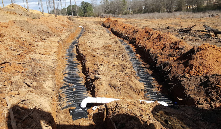Trenches with black corrugated drainage pipes, likely for a septic system, in a dirt field with trees in the background.