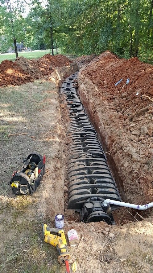 An open trench reveals a long, black, ribbed drainage system with tools scattered nearby in an outdoor setting.