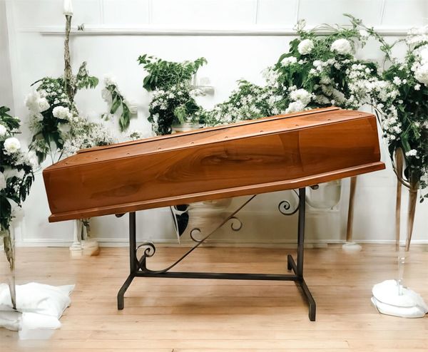 Una bara marrone chiusa, adornata con fiori, candele e steli di grano, contro una parete verde acqua.