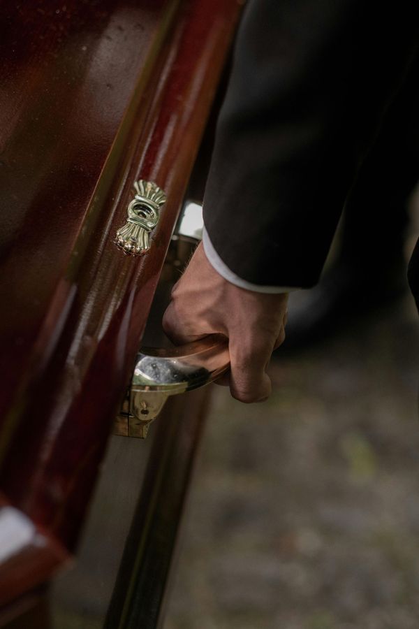 Mano di una persona sul manico di una bara, pronta a chiuderla. Legno scuro e dettagli dorati.