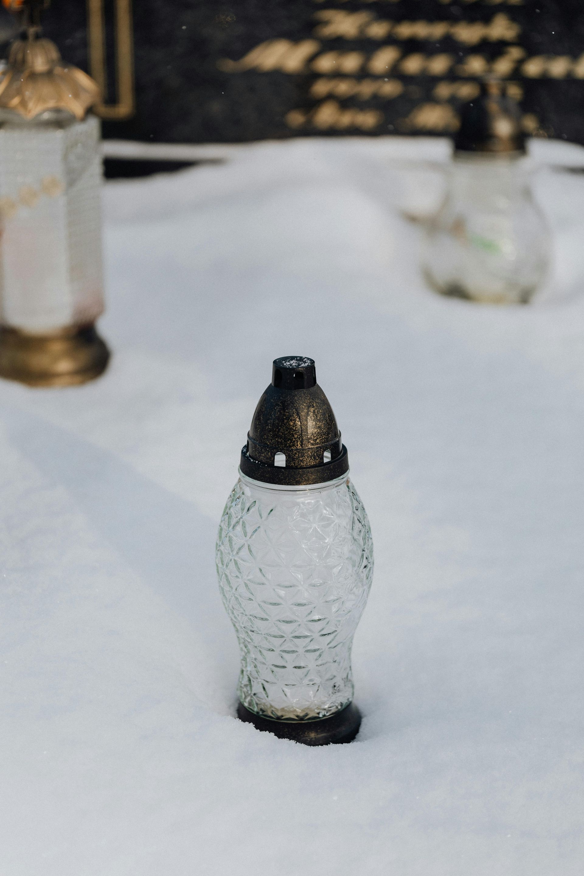 Lanterna con candela su terreno innevato in un cimitero, con una lapide scura visibile sullo sfondo.