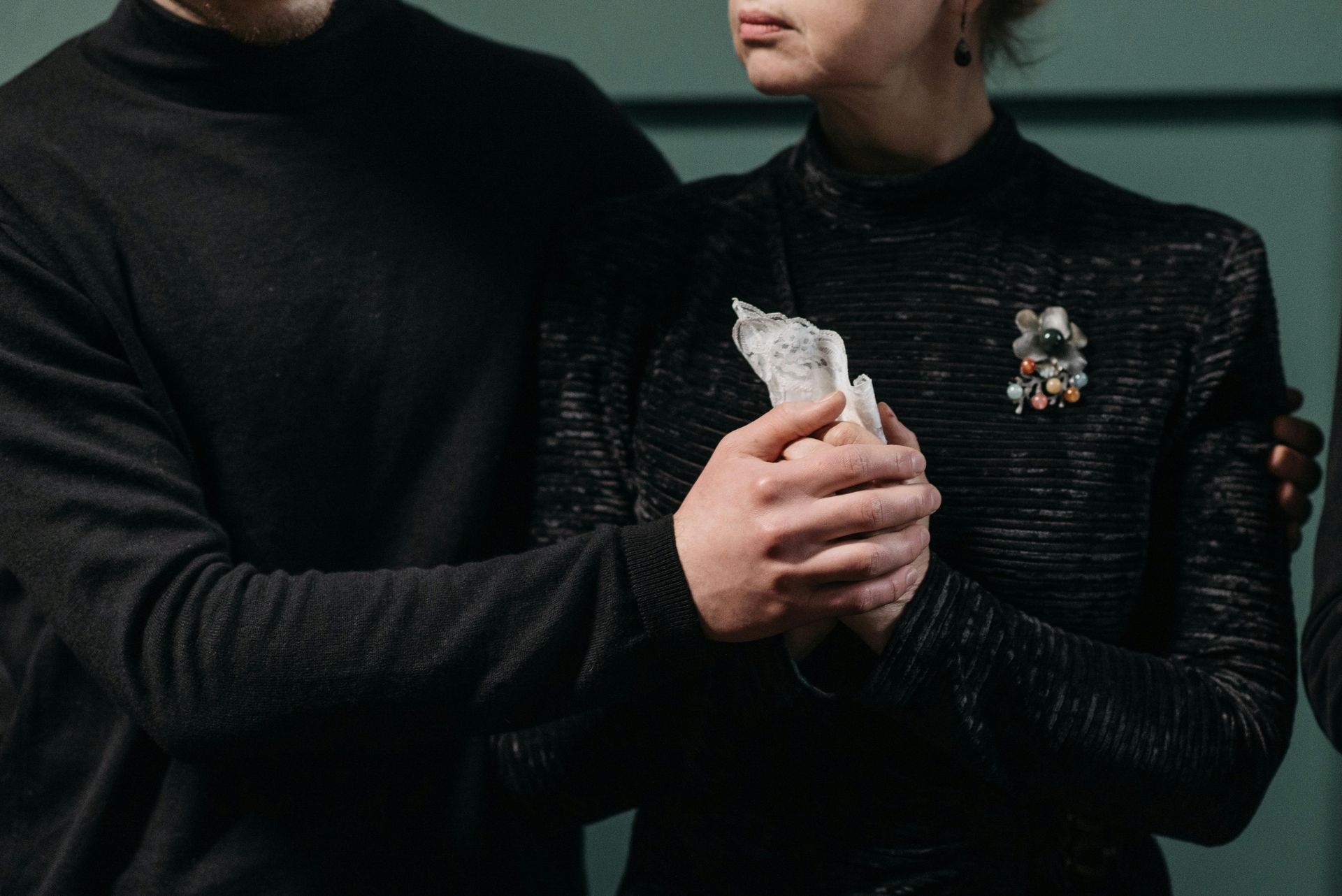 Una persona ne conforta un'altra; entrambe vestono di nero, la donna tiene in mano dei fazzoletti, la spilla è visibile.