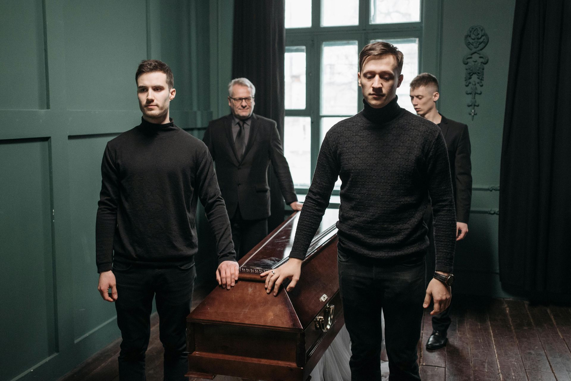 Quattro persone, una delle quali in giacca e cravatta, trasportano una bara in una stanza con pareti verdi e una finestra.