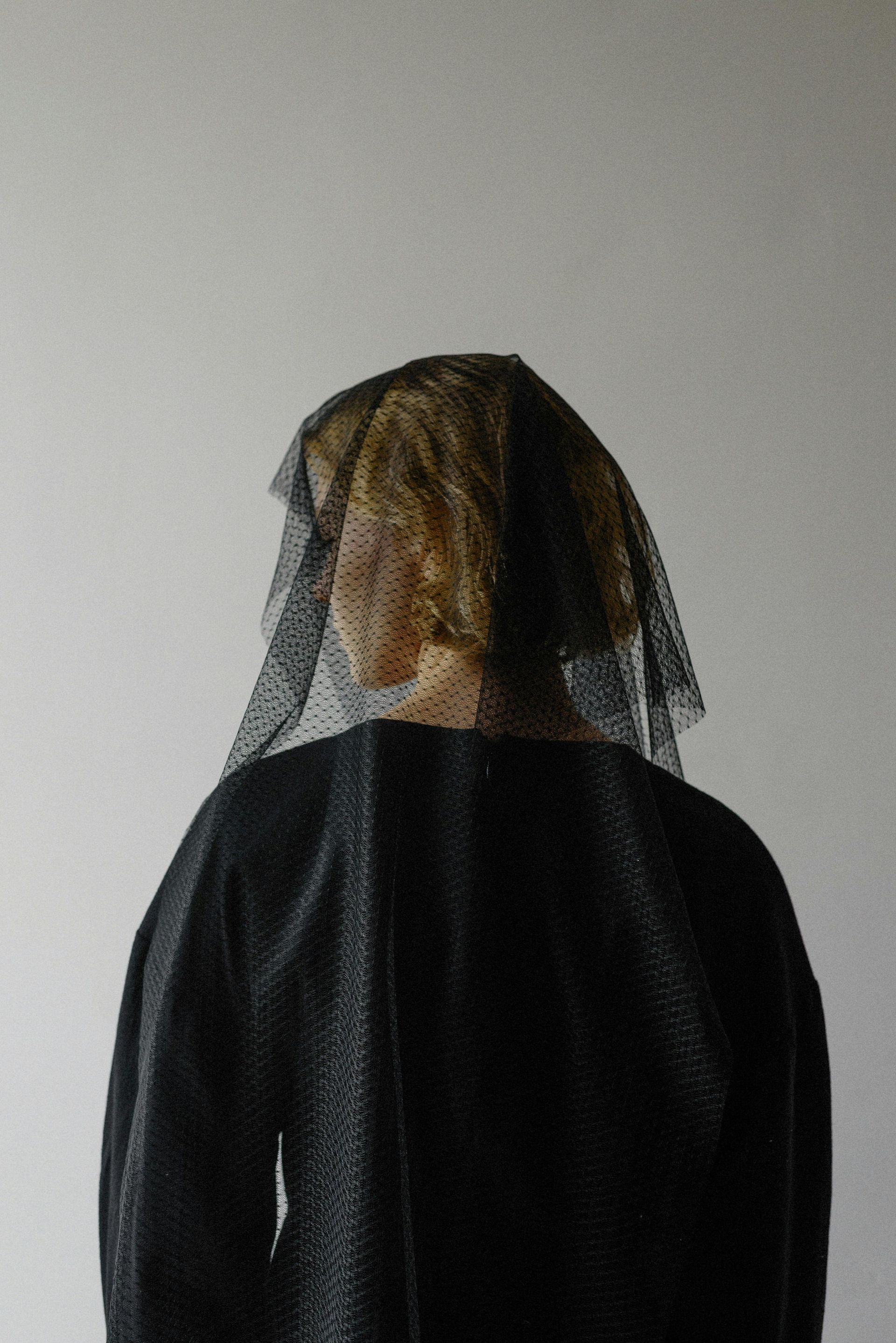 Persona vestita con abiti neri testurizzati, con la testa coperta da un velo nero trasparente, in piedi contro un muro bianco.