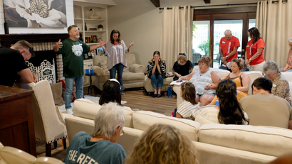A group of people gathered inside a home; a man speaks with arms open wide.