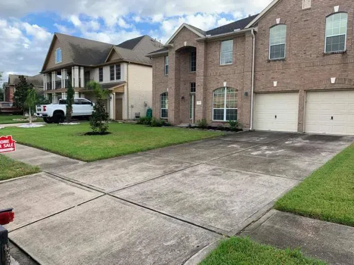 A residential street view featuring a tan brick house with a multi-car driveway next to a white house with a green lawn.