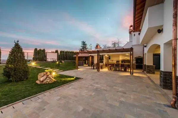 A paved patio area with an open-air wooden pergola dining space next to a modern house at sunset.