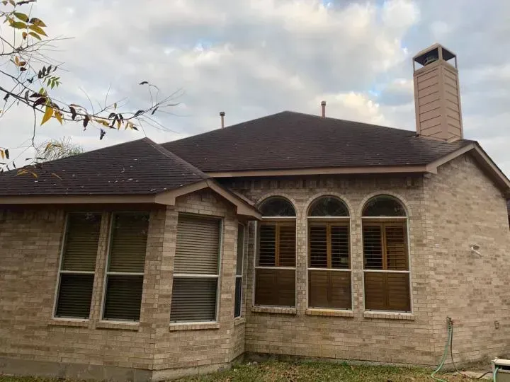 A tan brick house with a dark shingled roof, a chimney, and three arched windows on an overcast day.