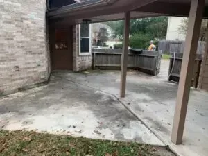 A concrete patio under a covered entryway leads to a wooden fence and grassy area outside a brick building.