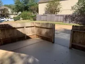 A wooden fence surrounds a gray concrete patio, opening to a residential driveway and neighboring house.