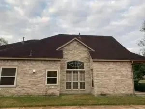 A tan brick house with a dark brown shingled roof, featuring a central arched window under a peaked gable.