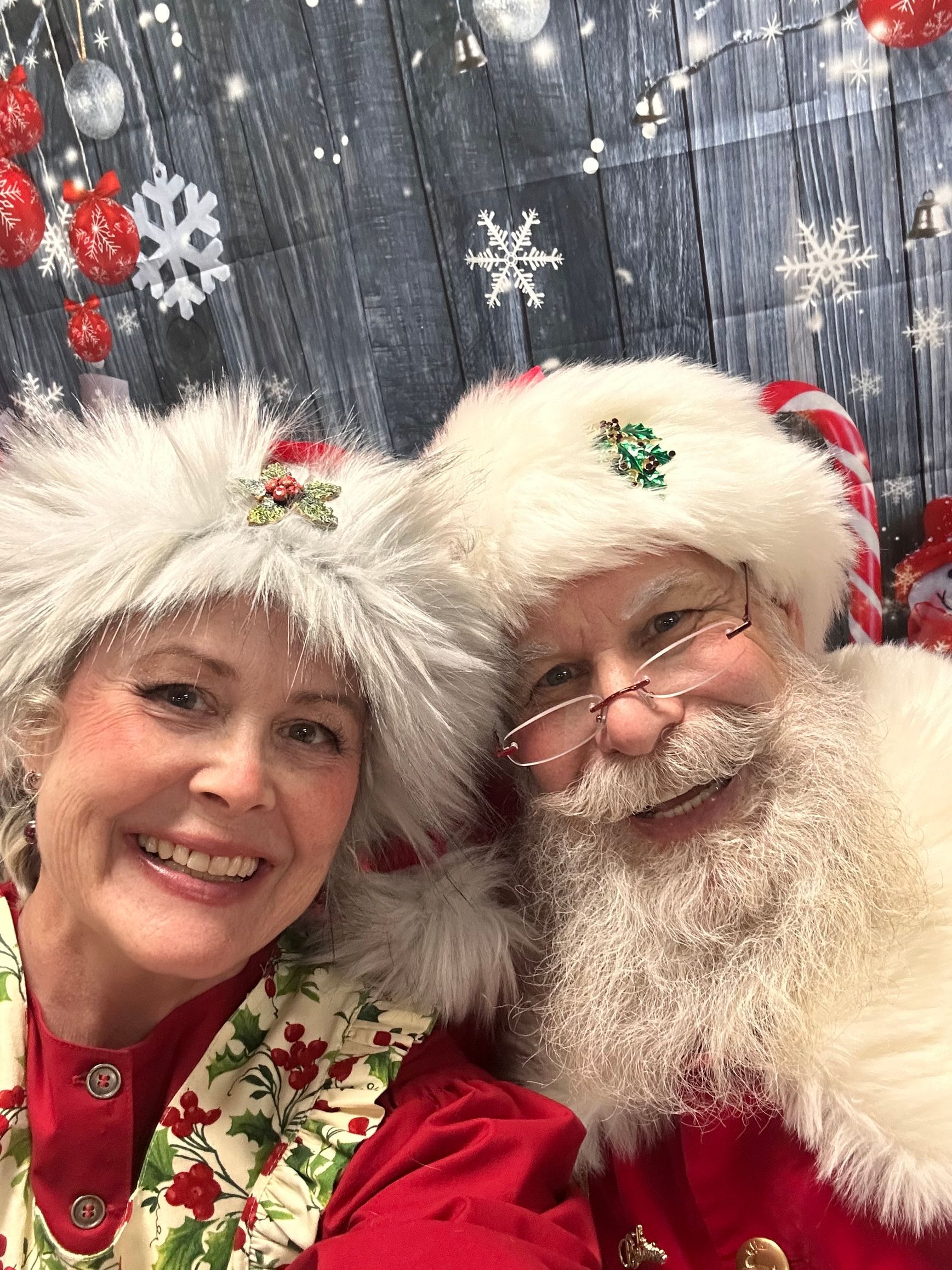 Santa and Mrs. Claus smiling close-up, wearing red and white, in front of a Christmas background.
