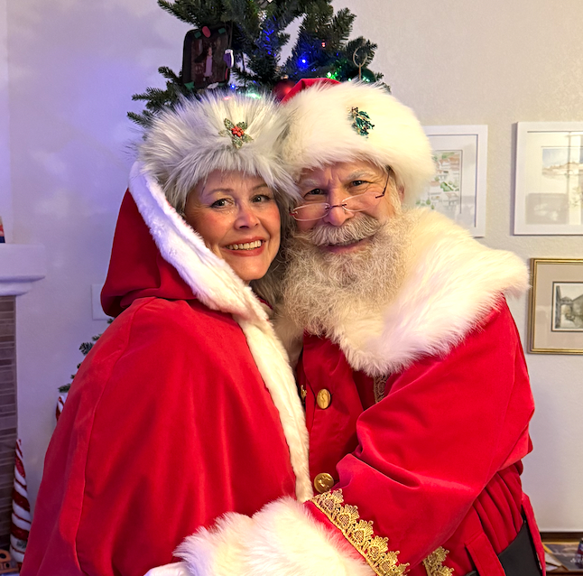 Santa Claus and Mrs. Claus hugging in front of a Christmas tree, smiling, wearing red and white festive attire.