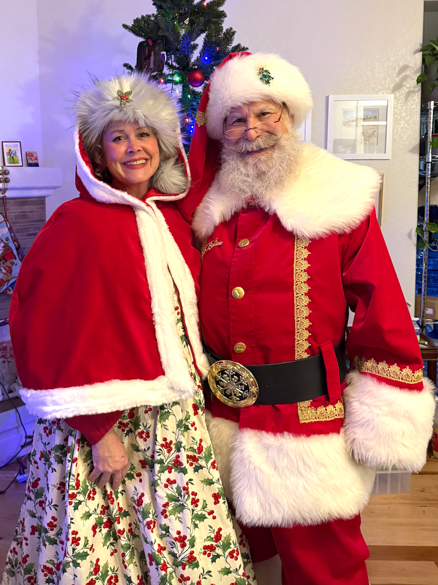Santa and Mrs. Claus smiling, posing for a photo by a Christmas tree.