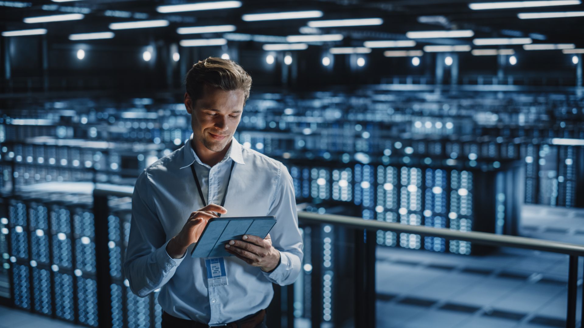 A man is standing in a server room looking at a tablet.