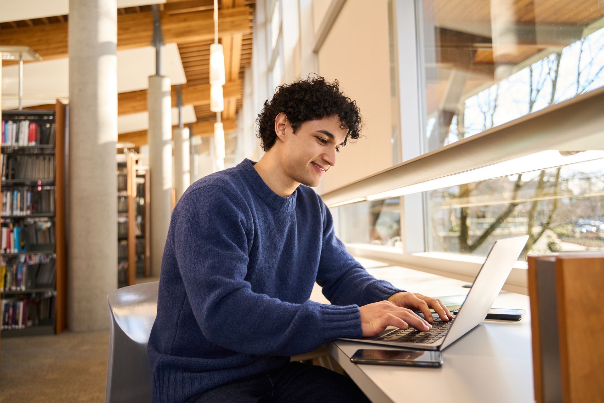 A man is sitting at a desk in a library using a laptop computer.