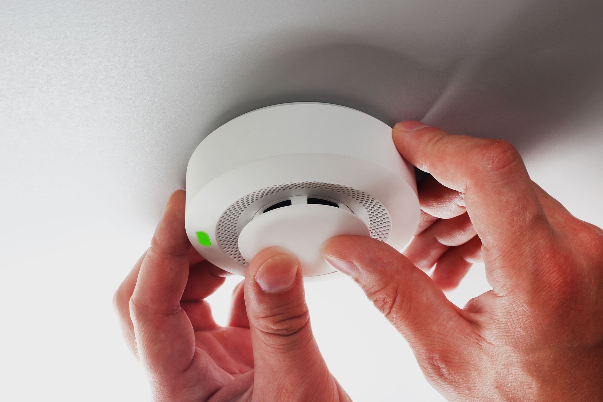 Hands installing a white smoke detector on a white ceiling.