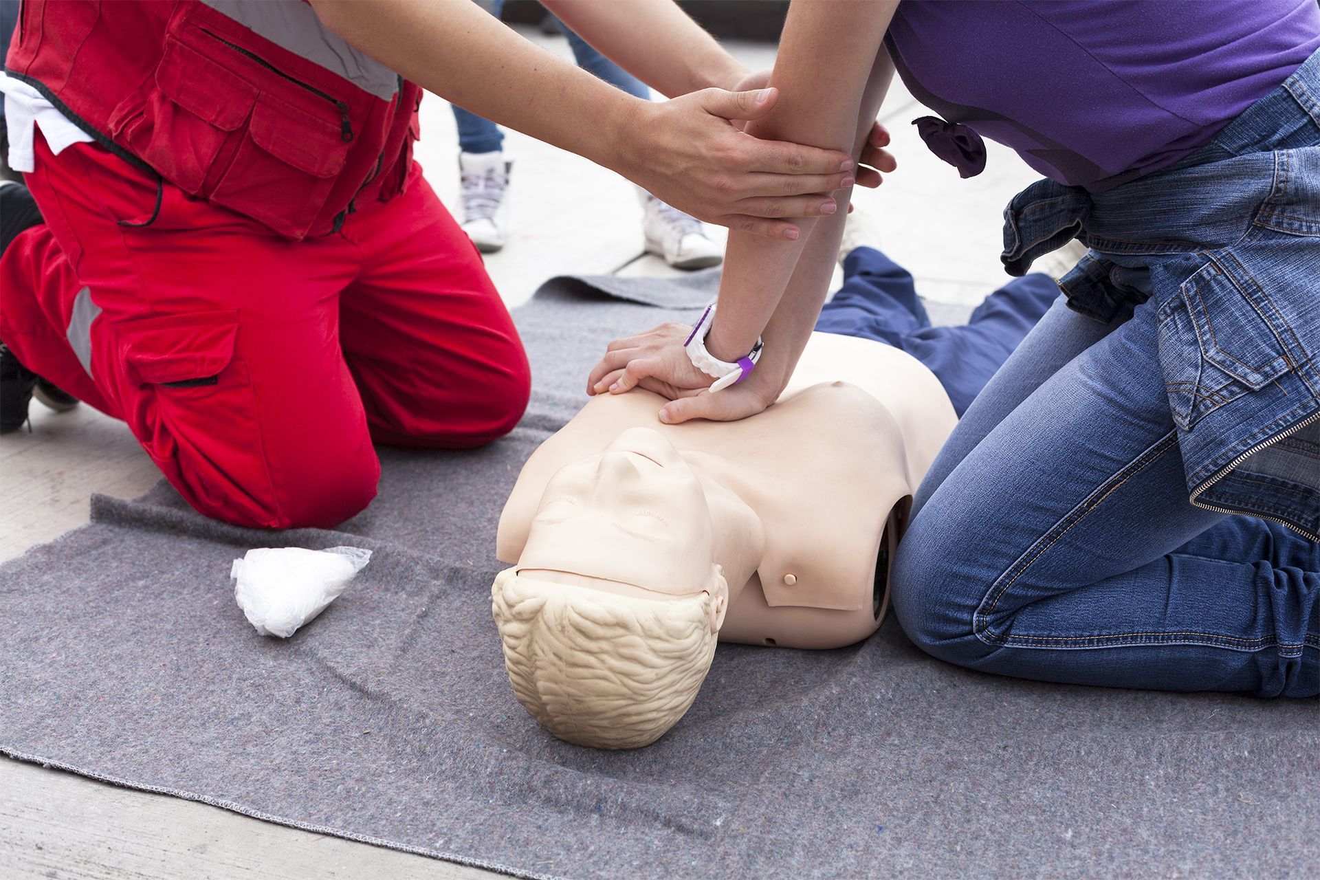 Two people practicing CPR on a dummy; one in red, one in blue jeans. Outdoors.
