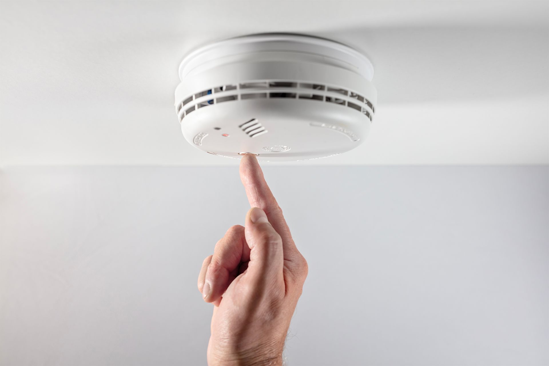 Hand pressing the test button on a white smoke detector mounted on a white ceiling.