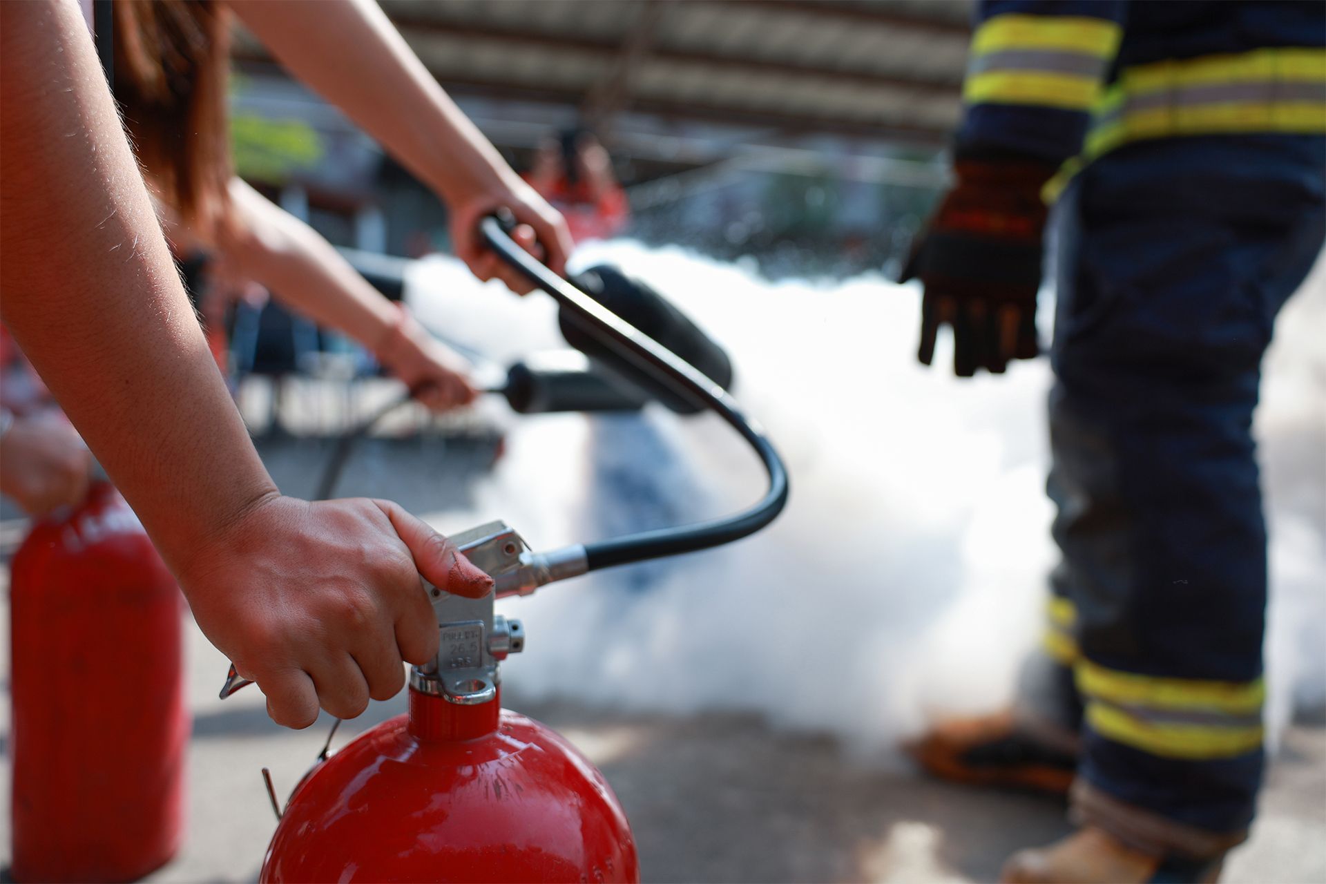 Hands operating a red fire extinguisher, releasing white foam. A firefighter stands nearby.
