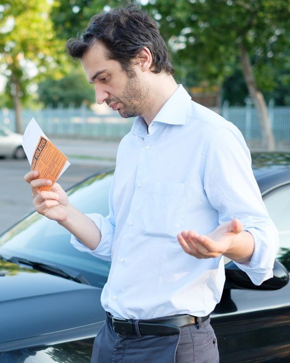 Un homme, l'air perplexe, regarde une contravention de stationnement, debout près de sa voiture noire.