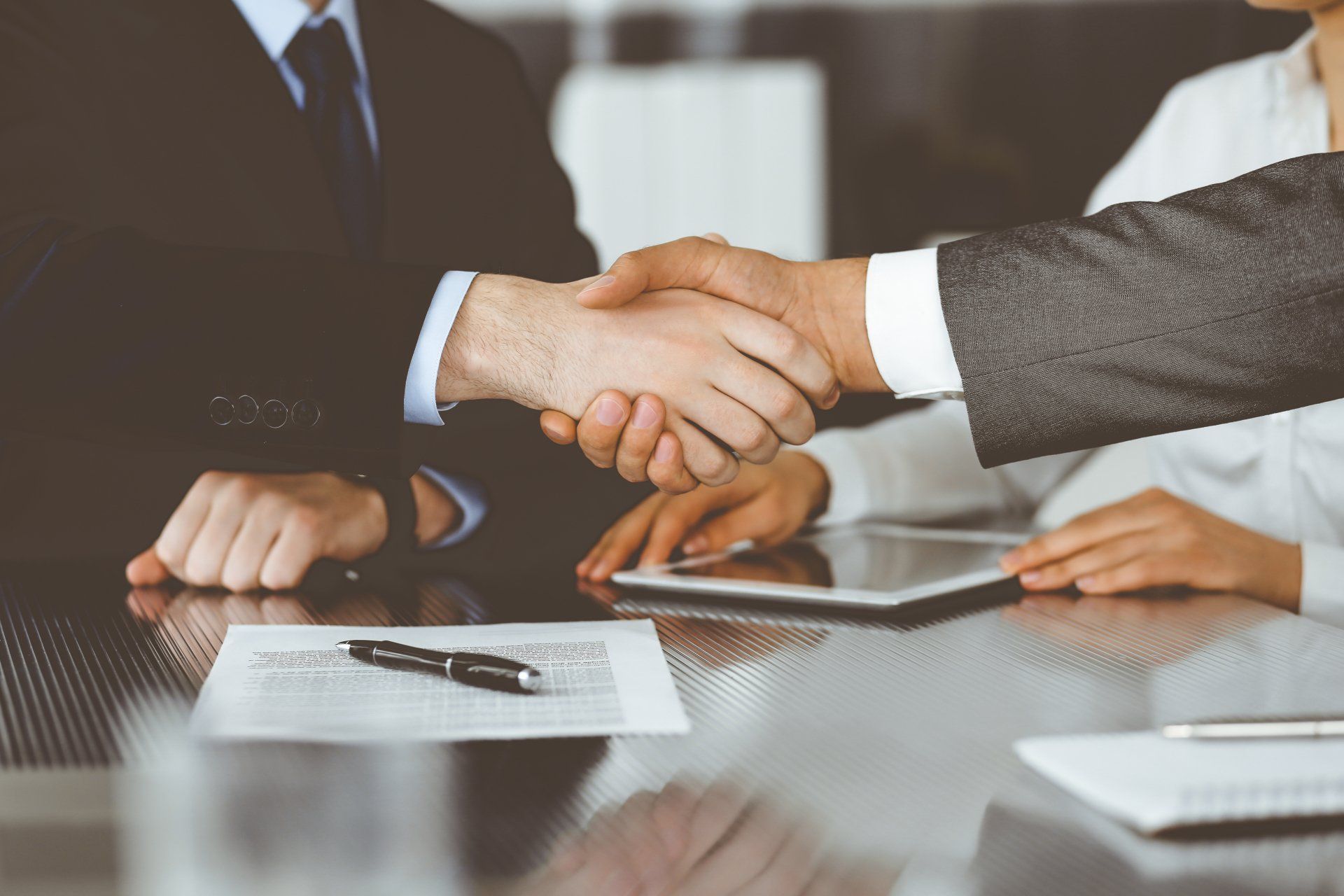 Deux hommes en costume se serrent la main au-dessus d'une table où se trouvent des documents et une tablette, symbolisant une transaction commerciale.