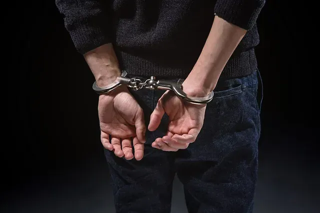 Personne menottée, vue de dos, portant un jean et un pull noir sur fond sombre.