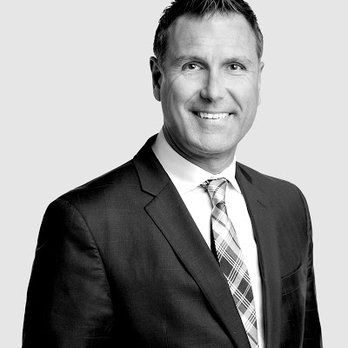 Homme en costume souriant, portant une cravate à motifs sur un fond gris.