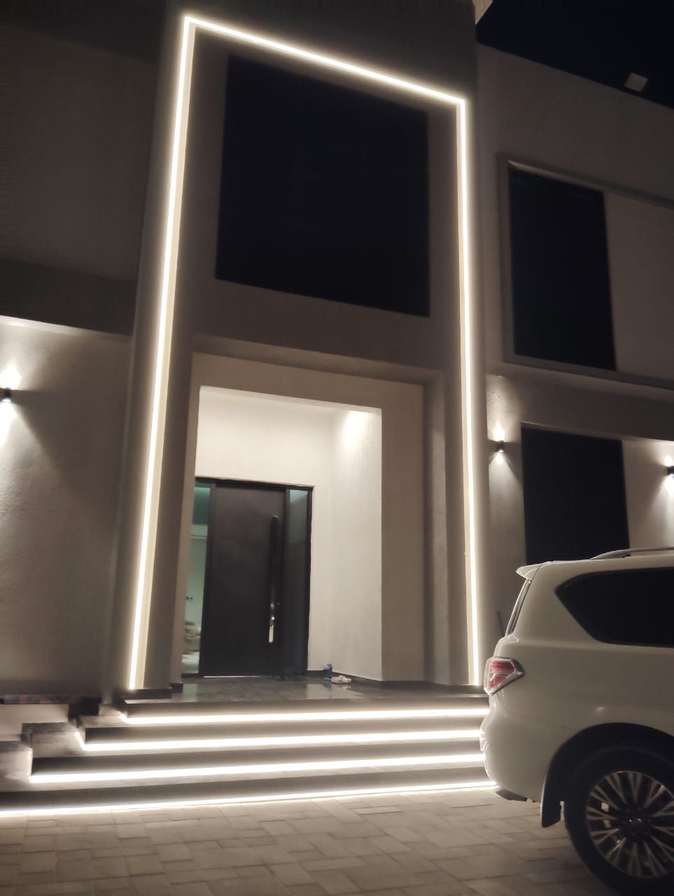 Modern home exterior at night, white with illuminated accents. SUV parked.