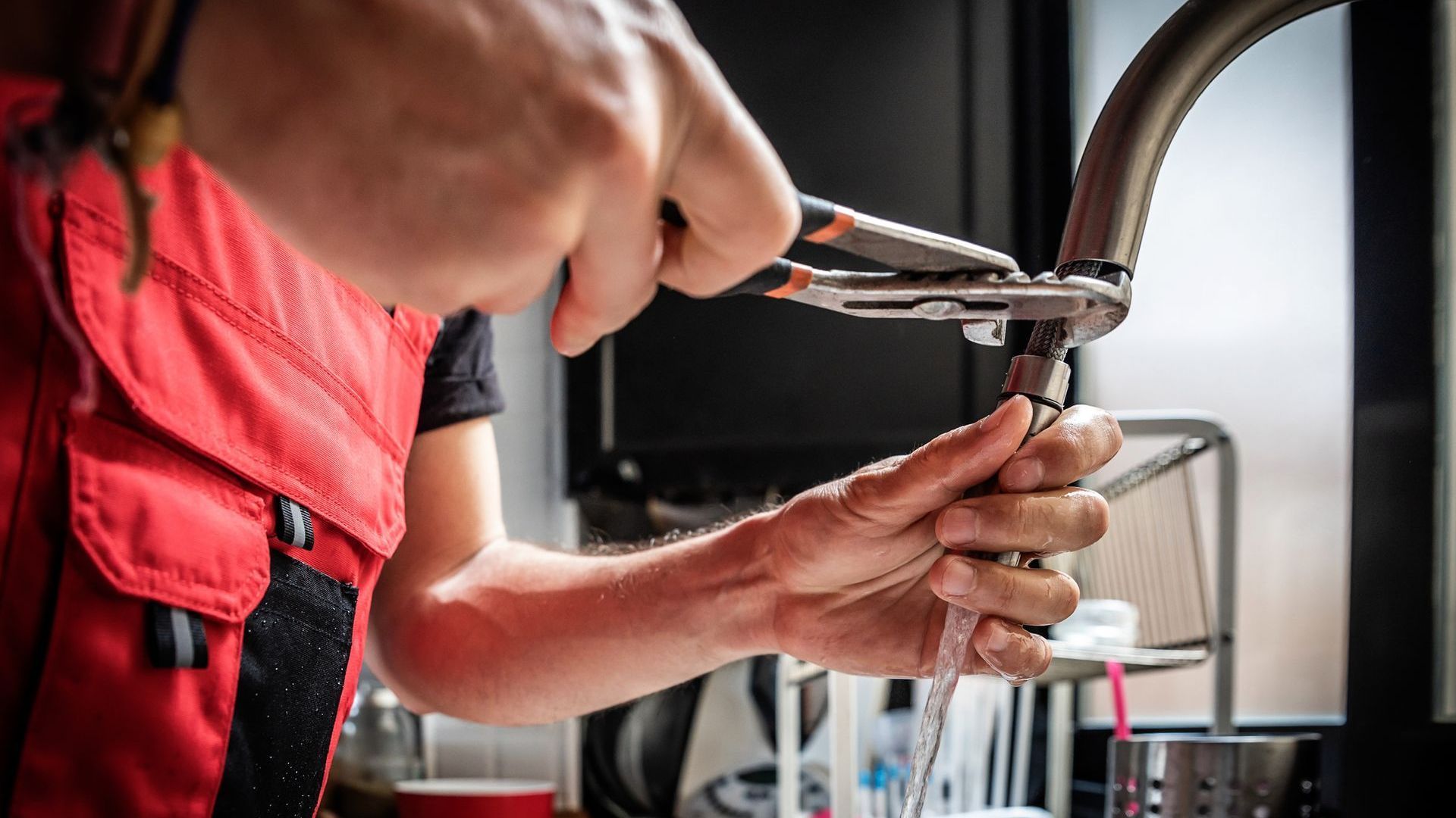 A person in a red vest repairs a kitchen faucet with pliers, water running.