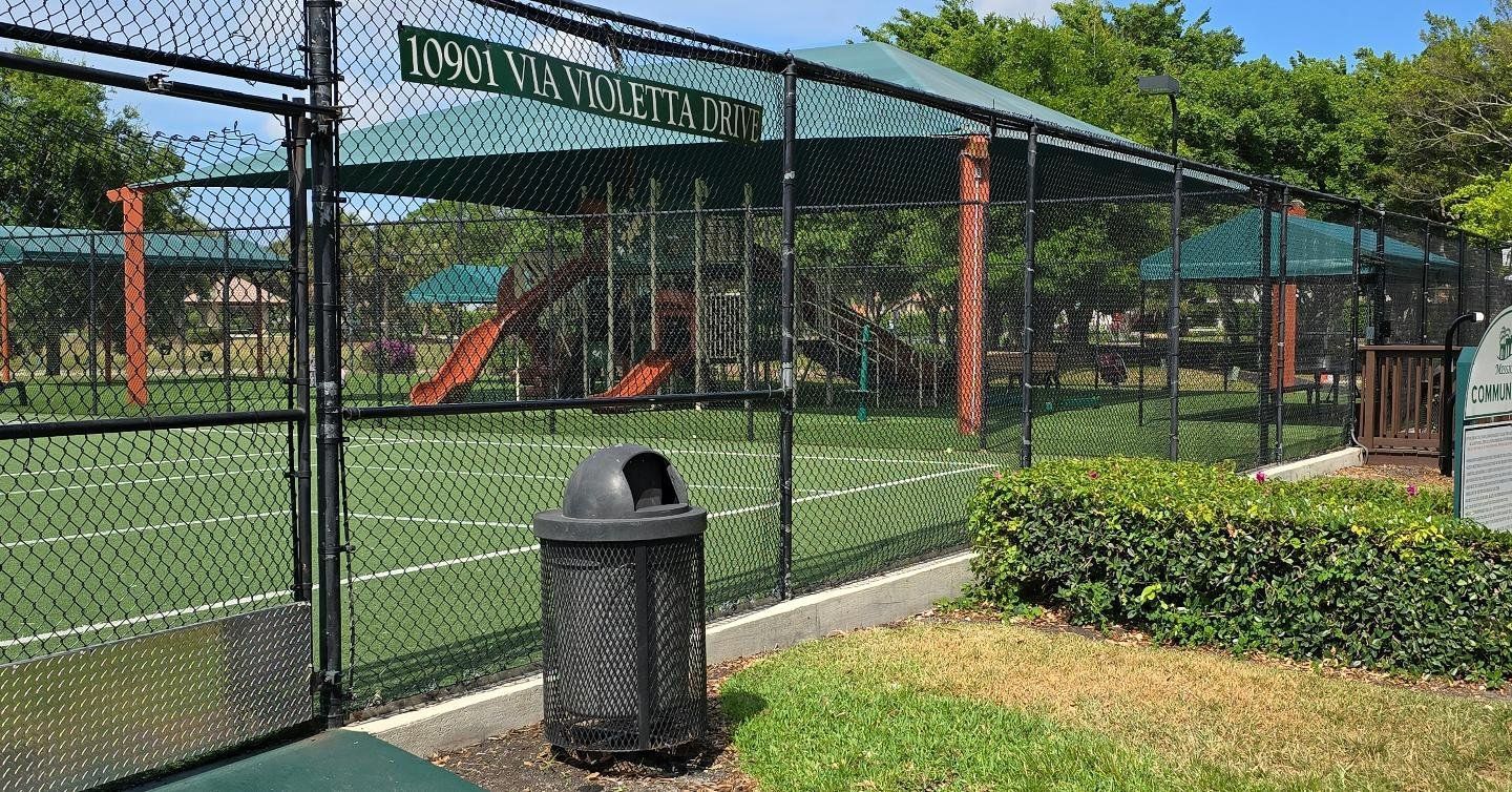 A trash can is sitting in front of a chain link fence in a park.