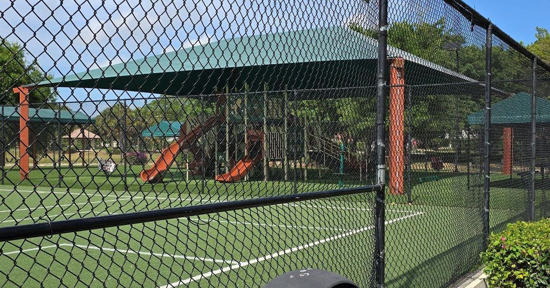 A chain link fence surrounds a tennis court in a park.