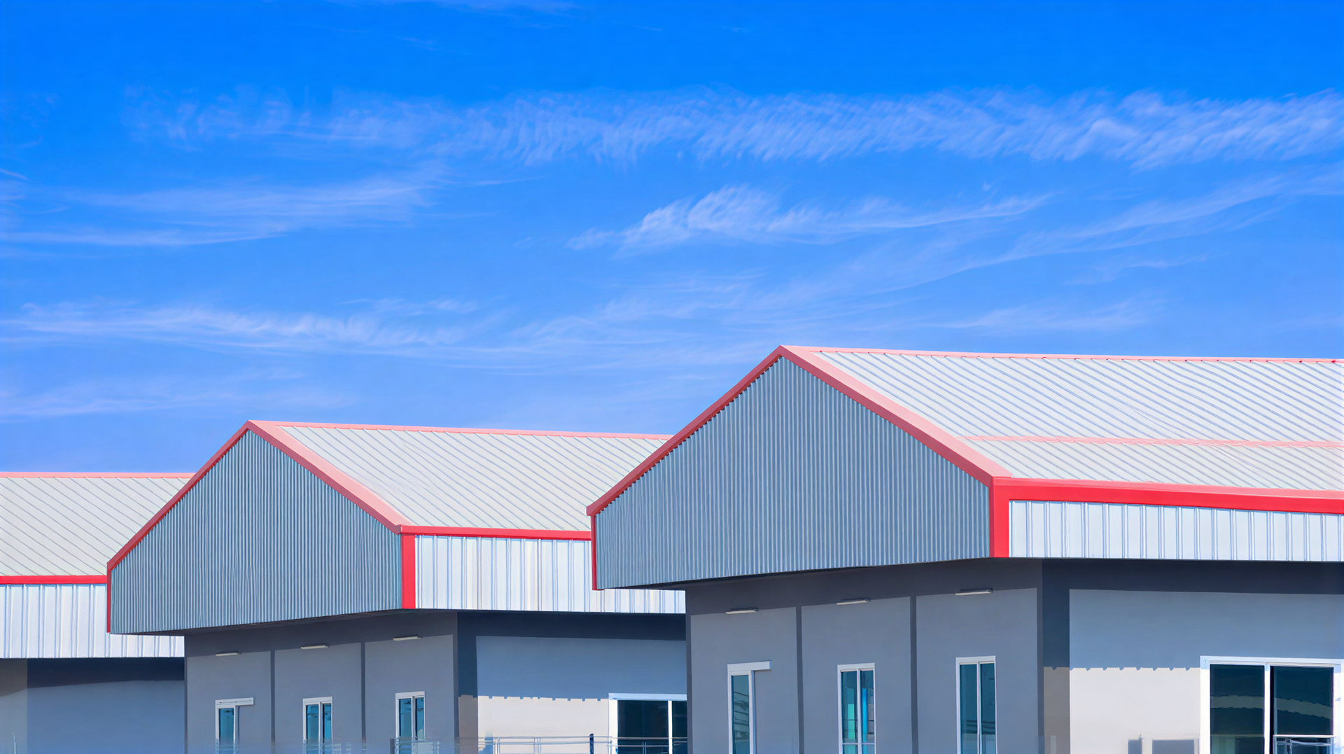 Row of gray industrial buildings with red-trimmed, angled metal roofs against a bright blue sky.