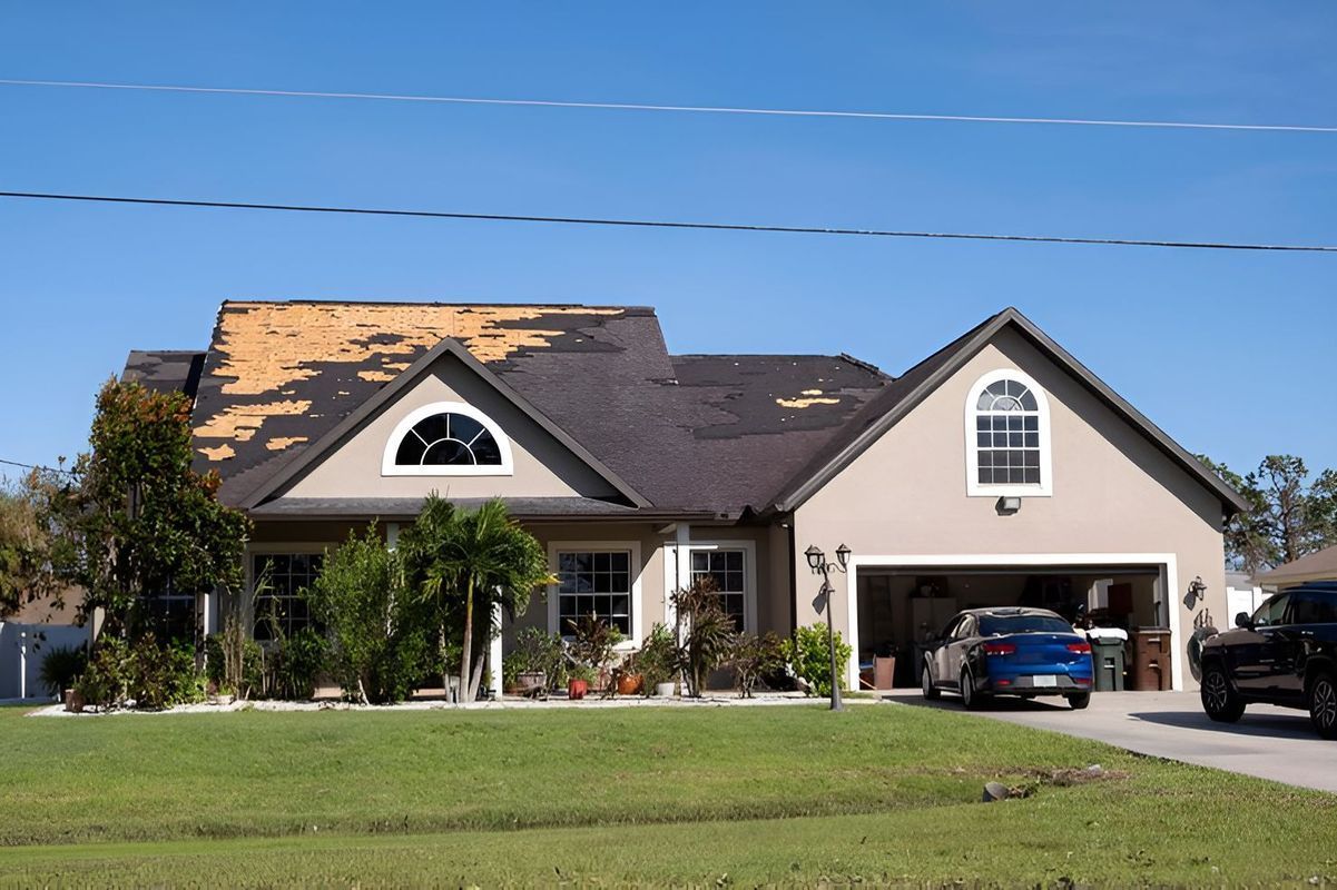 House with damaged roof and cars in the driveway under a clear blue sky.