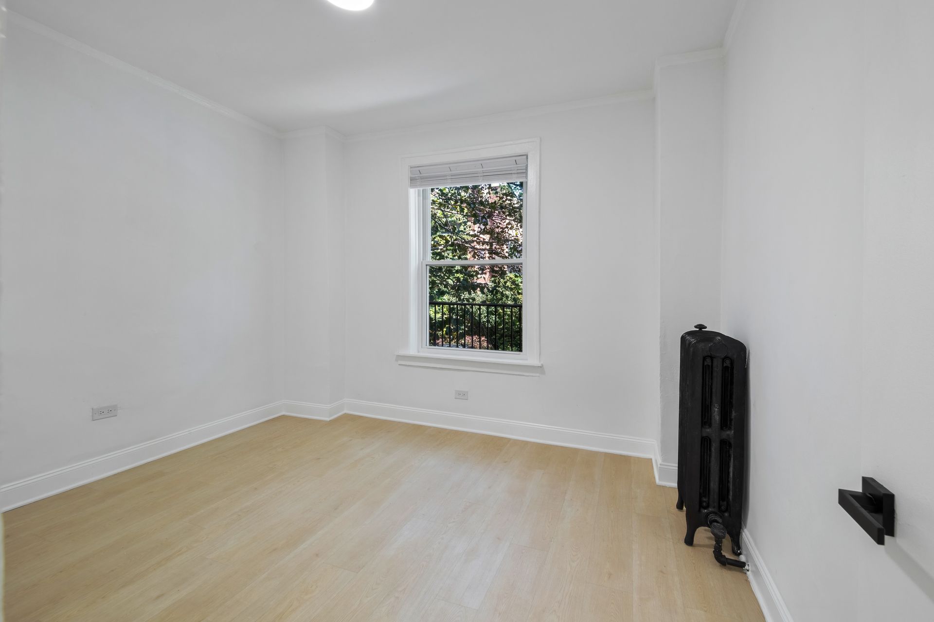 Empty white room with window, light wood floor, and black radiator.