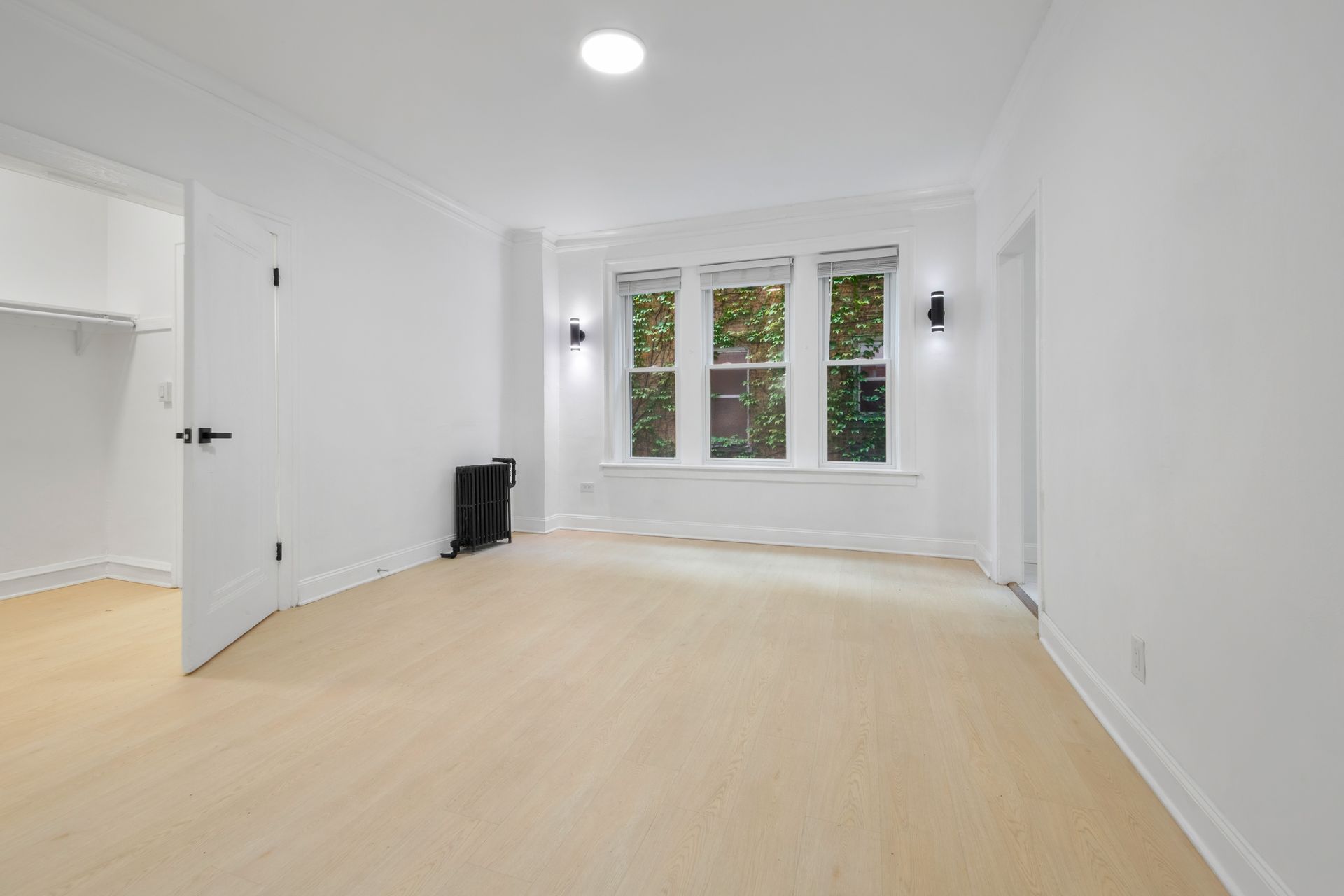 Empty white room with light wood floor, large window, open closet, and black radiator.
