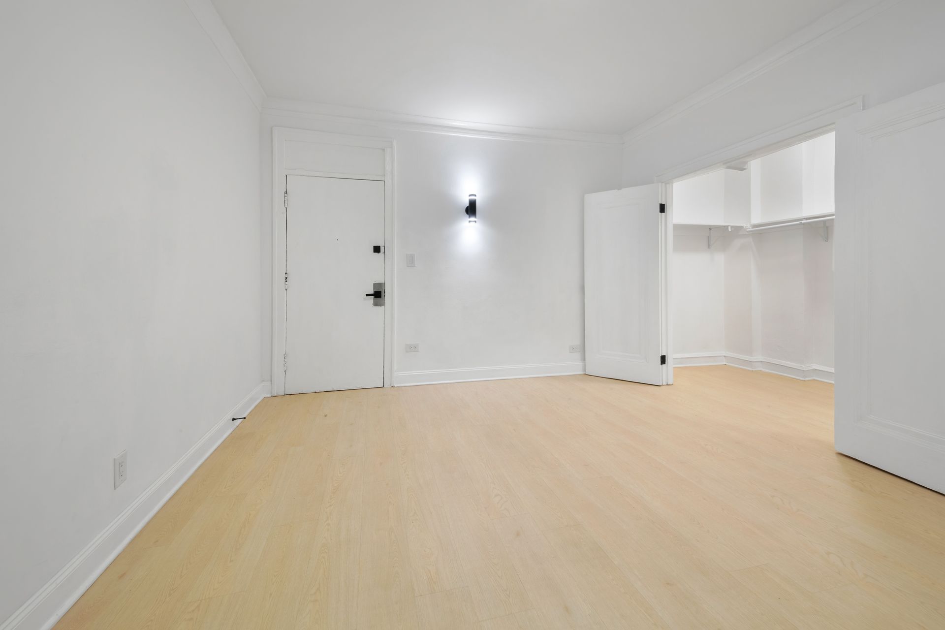 Empty, white-walled room with light wood floor. Open closet on right. Door on left.