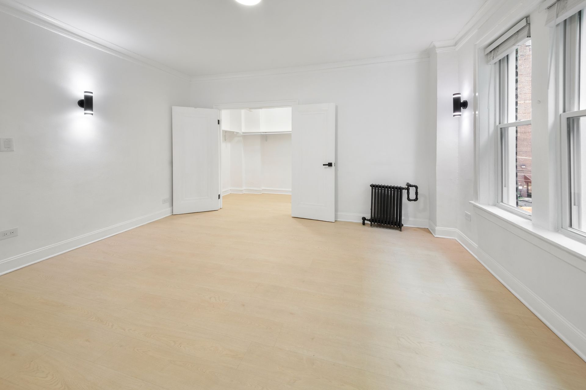 Empty white-walled room with wooden floors, doors to a closet, and windows. Black wall lights and radiator.