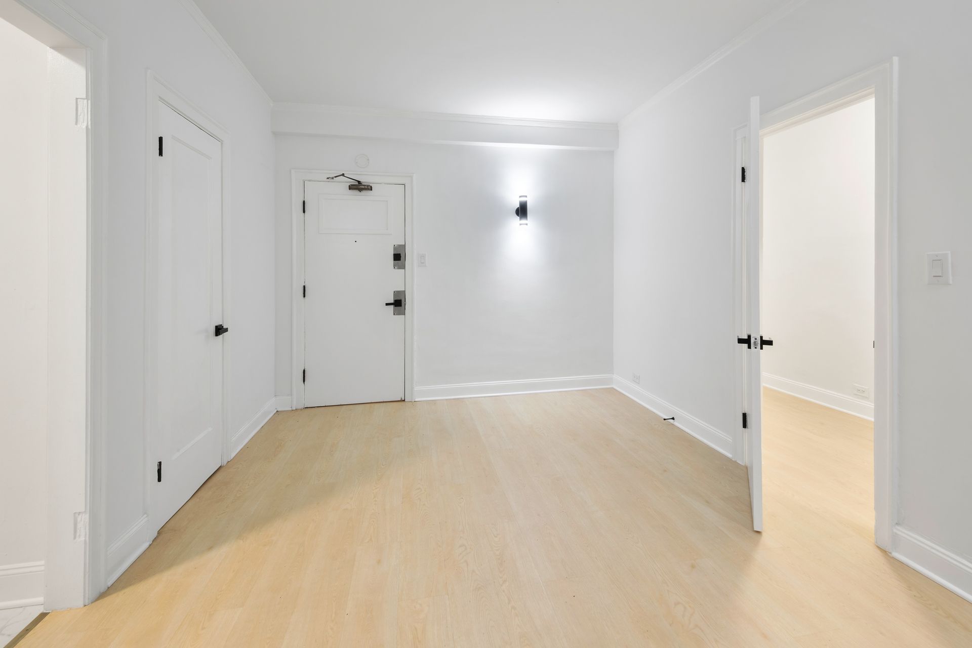 Empty, bright white hallway with light wood flooring, three white doors, and sconce lighting.