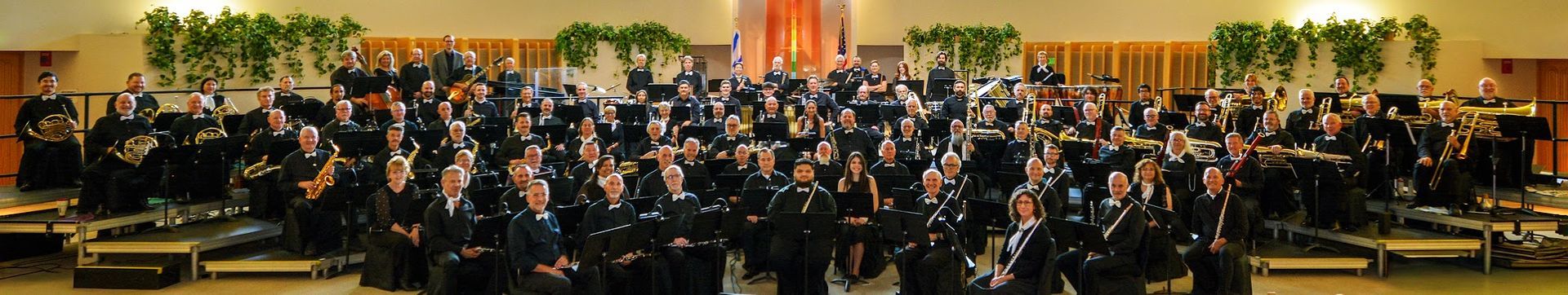 This is a photograph of the entire 110 member symphonic band dressed all in black with white ties and scarves.  The band is sitting on risers on the stage of Temple Isaiah/Jewish Community Center in Palm Springs before our 2026 Spring Concert.