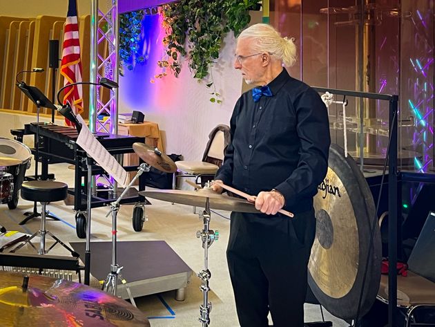 A man with white hair plays a cymbal in a concert setting. He stands near a gong and percussion instruments, wearing a black shirt and bow tie.