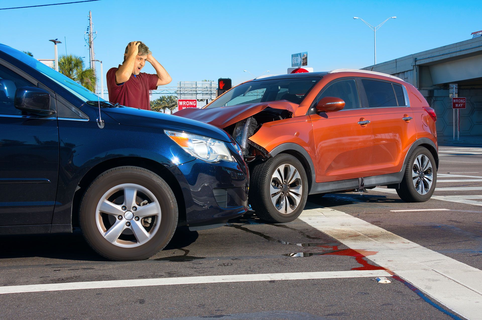 Two cars have crashed wrecked into each other at intersection with very upset man driver looking at the severe damage with wrong way sign in background.