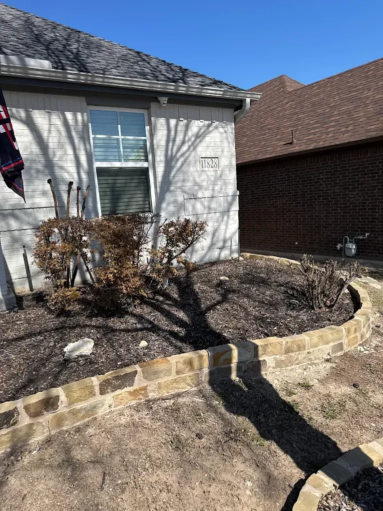 Front of a house with a raised garden bed and dead bushes under a sunny sky.