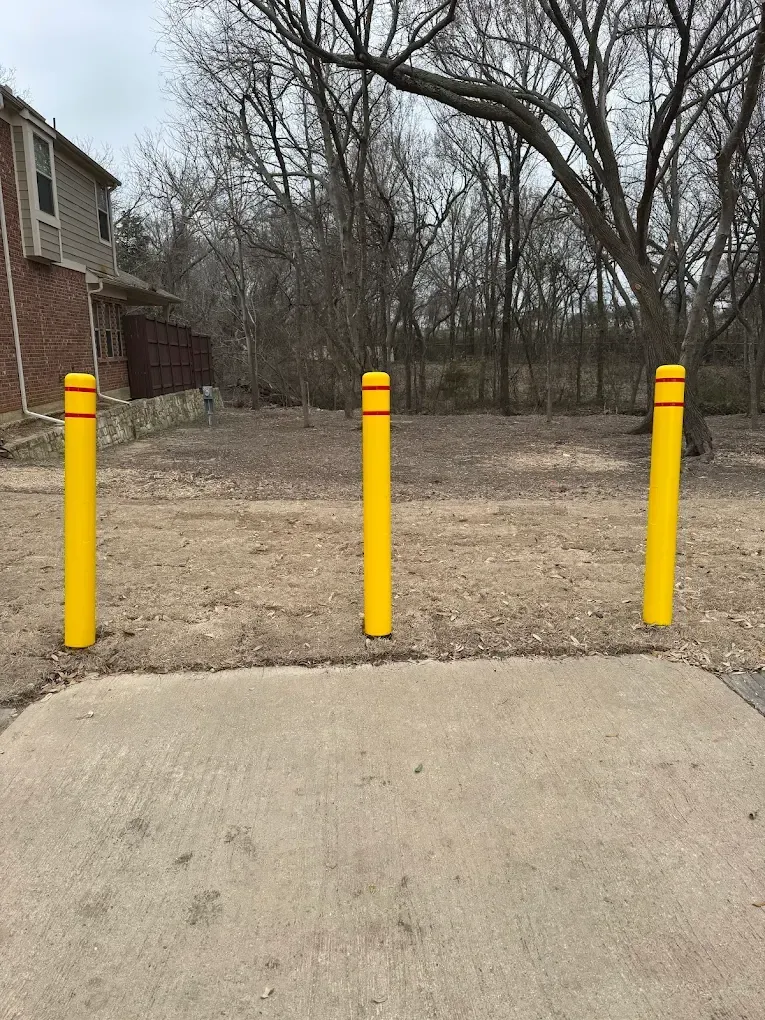 Three yellow bollards with red bands stand in front of trees and a building.