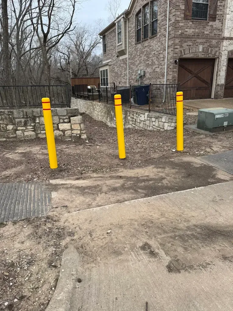 Three yellow bollards block a pathway. A brick building and retaining wall are in the background.