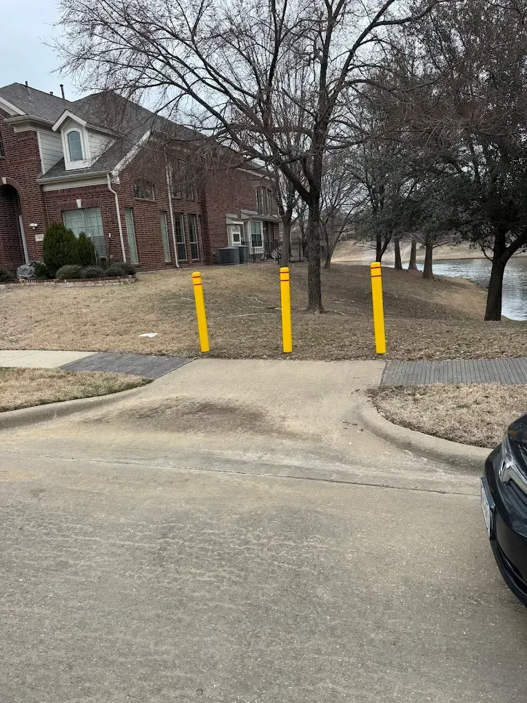 Yellow bollards block a sidewalk entry point, with houses and trees in the background.