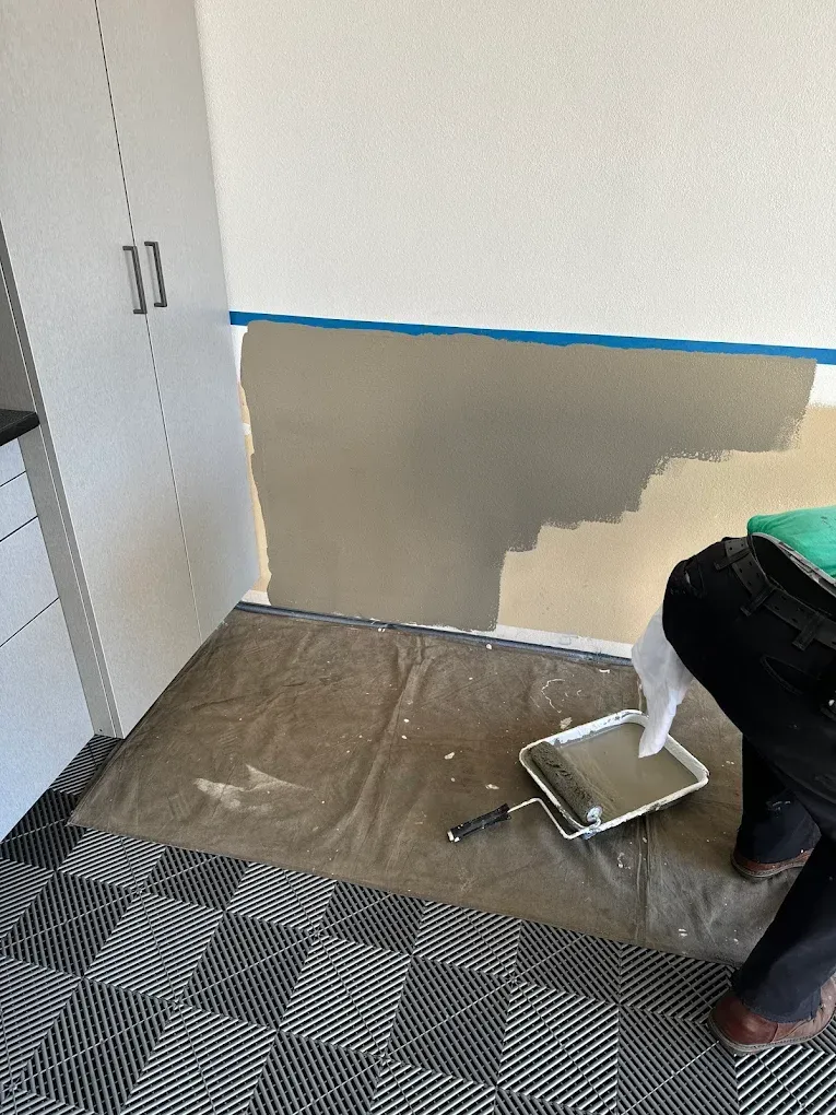 Person painting a wall, using a roller and tray. Room has cabinets, and checkered floor.