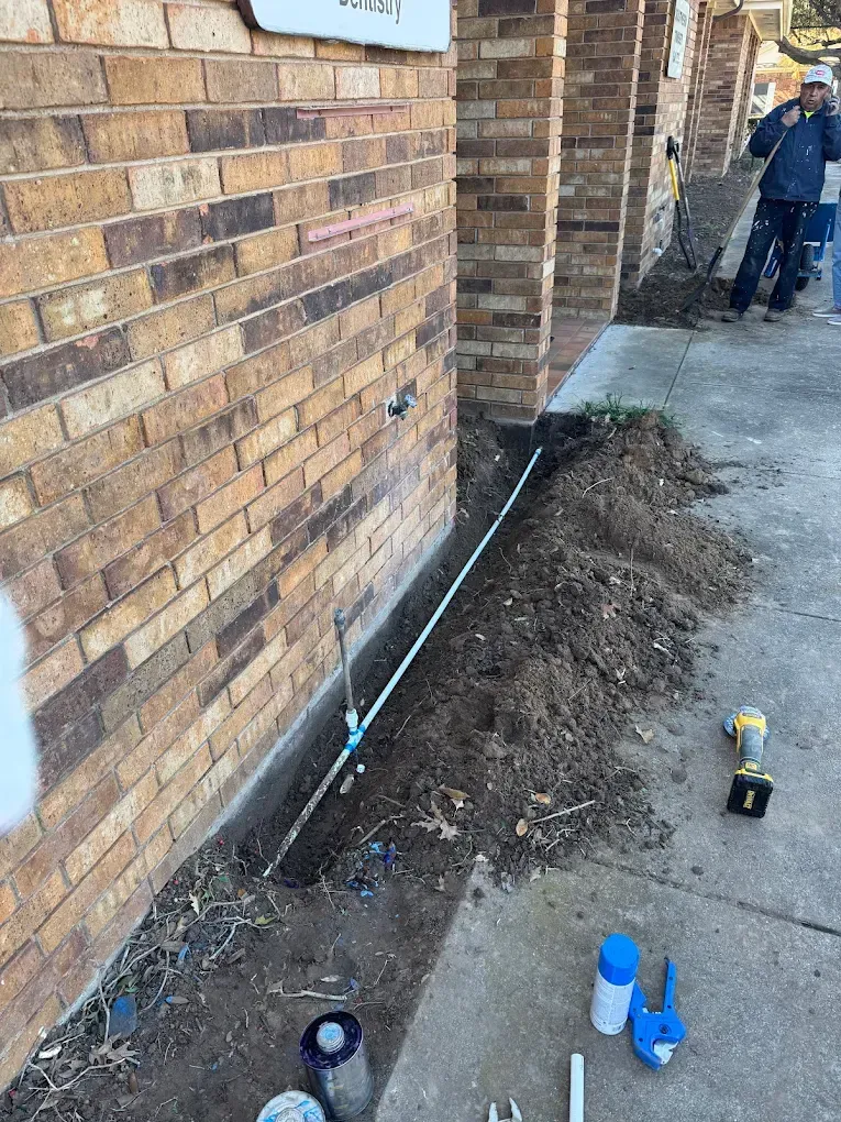 Construction workers working outside a brick building. Trench with white pipe. Ground is dug up.