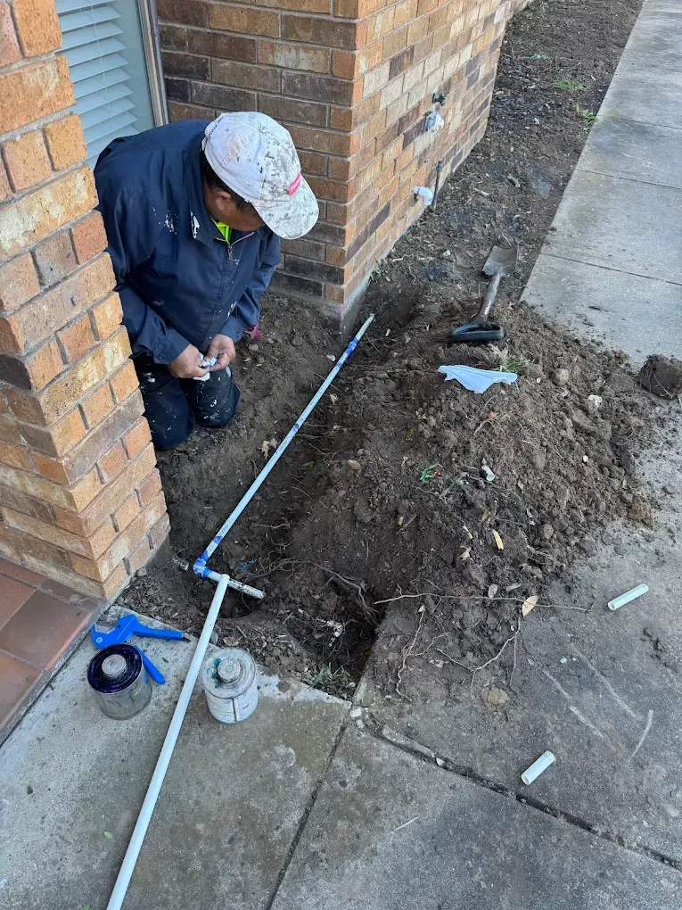 Man kneeling by brick building, working on irrigation pipes in a dirt trench, tools nearby.